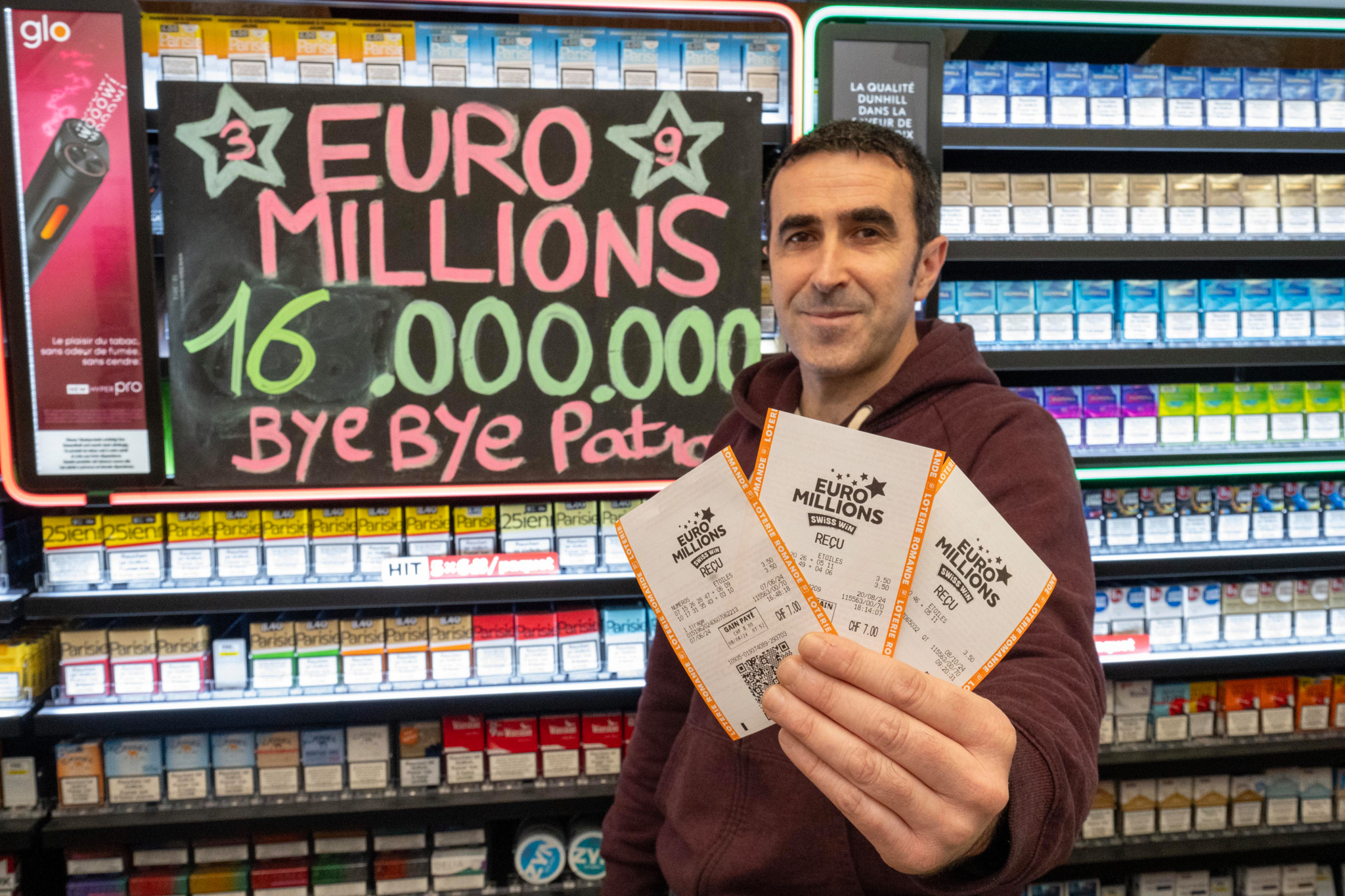 Un kiosquier à Lausanne tenant des billets Euro Millions devant une affiche indiquant 16 millions Bye Bye Patron.