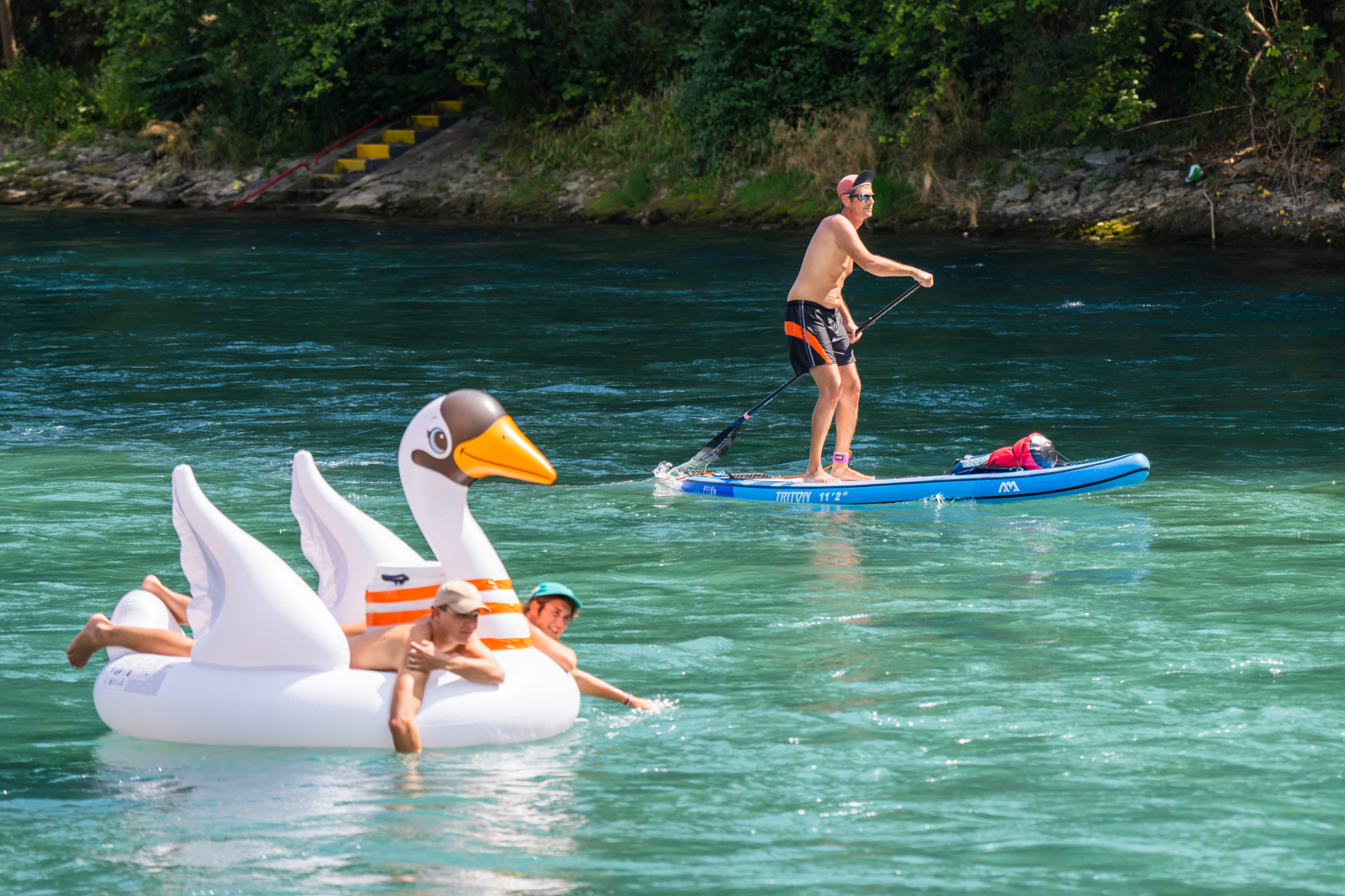 Kinder auf aufblasbarem Schwan-Schwimmtier und Mann auf Stand-up-Paddle-Board auf türkisblauem Fluss bei Hitze. Kinder auf aufblasbarem Schwan-Schwimmtier und Mann auf Stand-up-Paddle-Board auf türkisblauem Fluss bei Hitze.