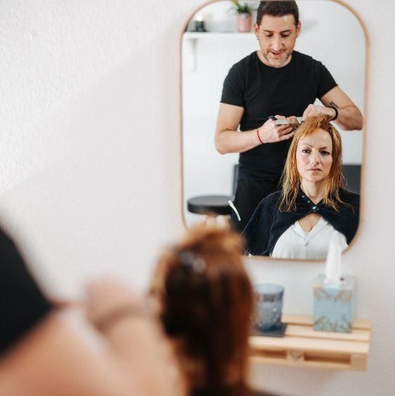 Un coiffeur coupe les cheveux d’une femme rousse assise devant un miroir, avec des outils de coiffure sur une étagère.