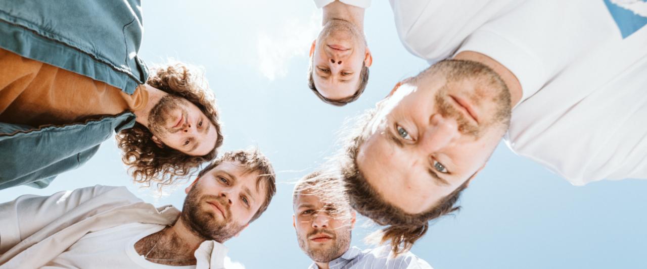 Fünf Männer blicken von oben in die Kamera, mit einem blauen Himmel im Hintergrund.