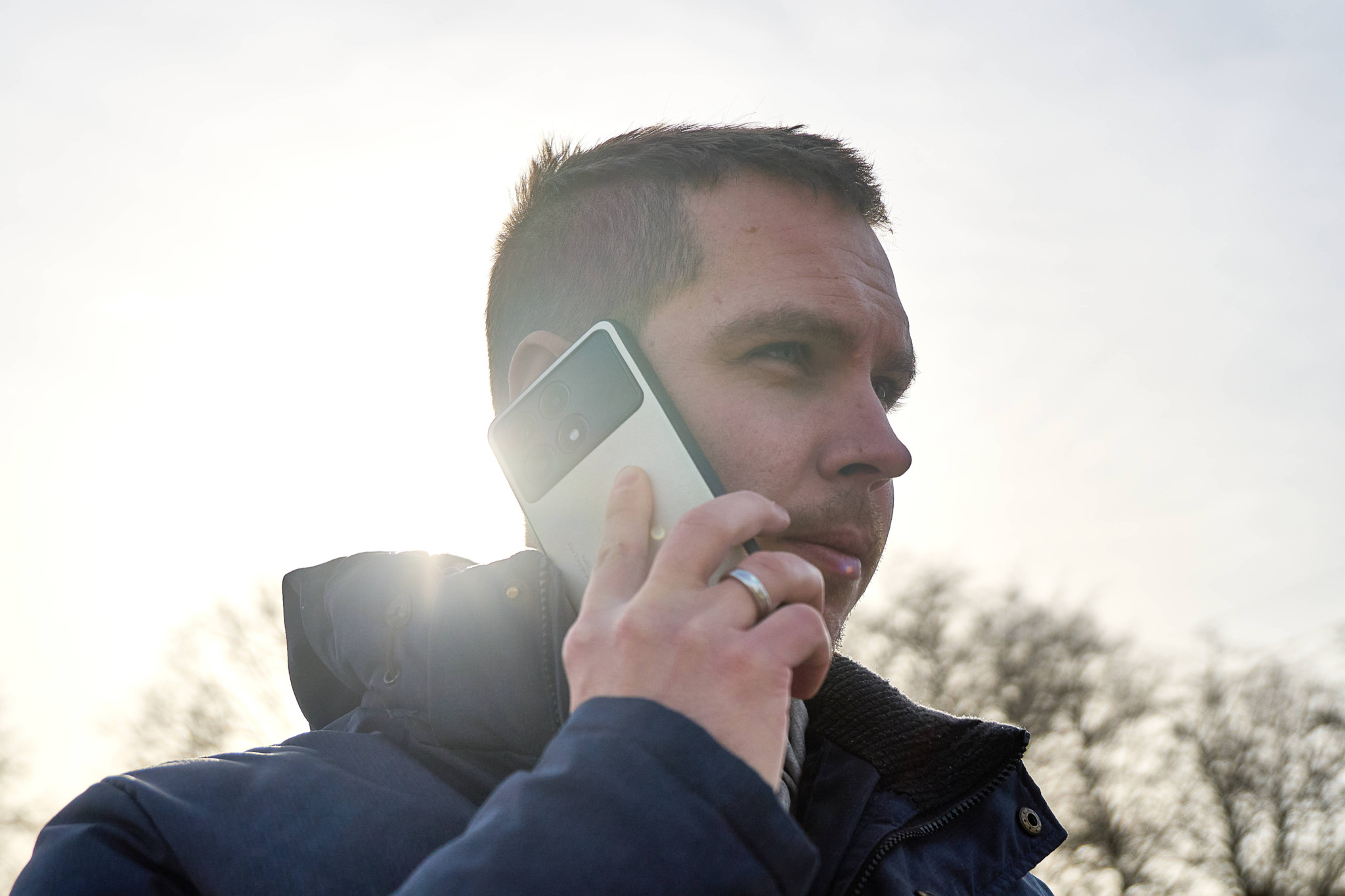 Un homme en veste bleue téléphone avec un smartphone dans un environnement urbain, ciel lumineux en arrière-plan.