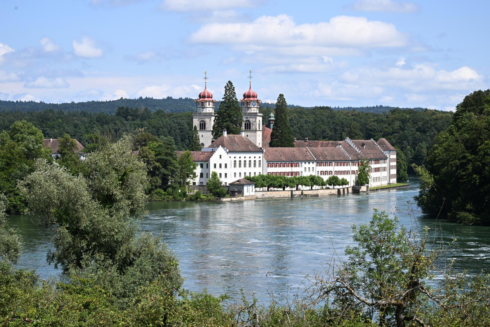 Luftaufnahme der Klosterinsel Rheinau, umgeben vom türkisblauen Rhein, mit Klostergebäuden und kleiner Kapelle inmitten üppiger Vegetation.