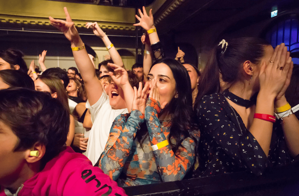 Des spectateurs enthousiastes applaudissent et crient lors d'un concert, certains portant des bracelets jaunes et des bouchons d'oreilles.
