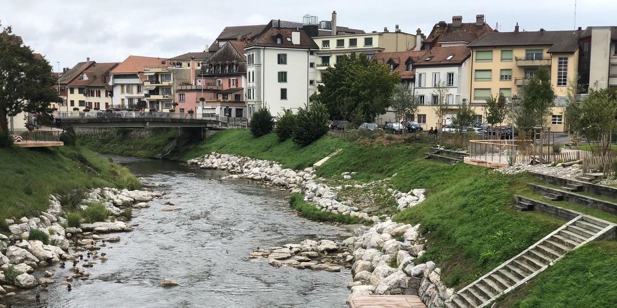 Vue sur une rivière traversant une ville avec des bâtiments colorés en arrière-plan, des rochers le long des berges et un pont en pierre traversant la rivière.