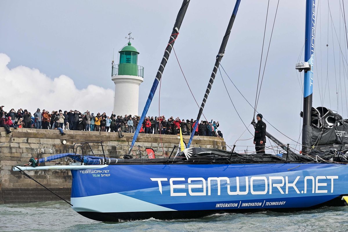 La skippeuse suisse Justine Mettraux se tient sur le pont du monocoque Imoca Teamwork.net en entrant dans le canal pour terminer la Vendée Globe en 8e position à Les Sables-d’Olonne, le 25 janvier 2025, avec un phare et des spectateurs en arrière-plan.