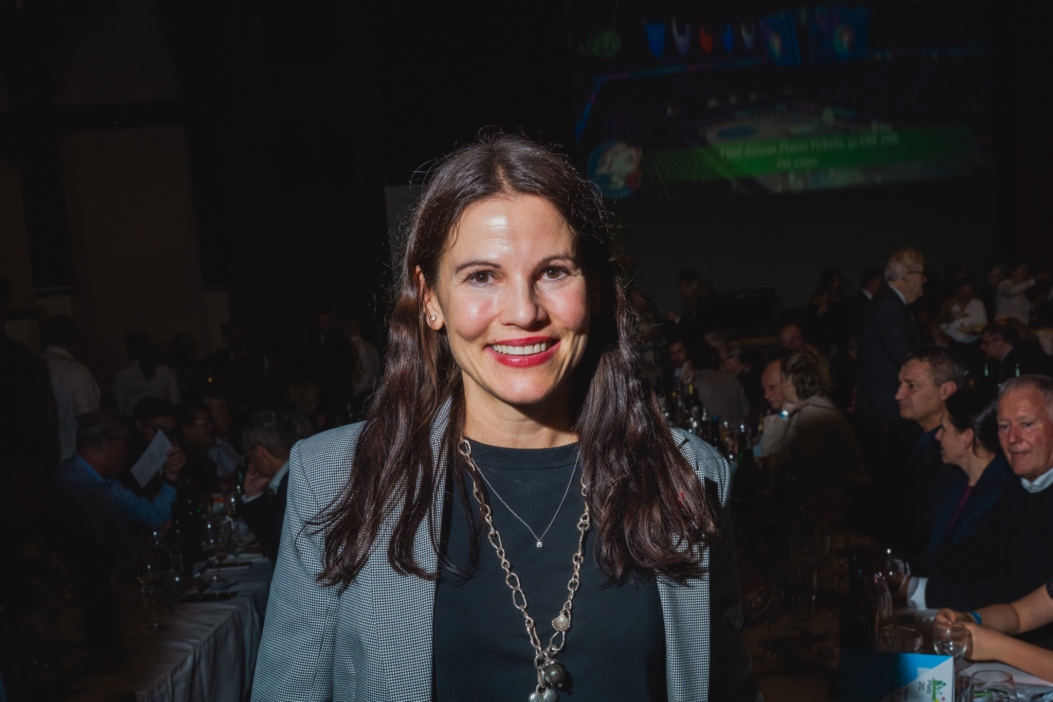 Frau mit dunklem Haar in einem grauen Blazer steht lächelnd in einem belebten Raum mit Tischen und Gästen im Hintergrund.