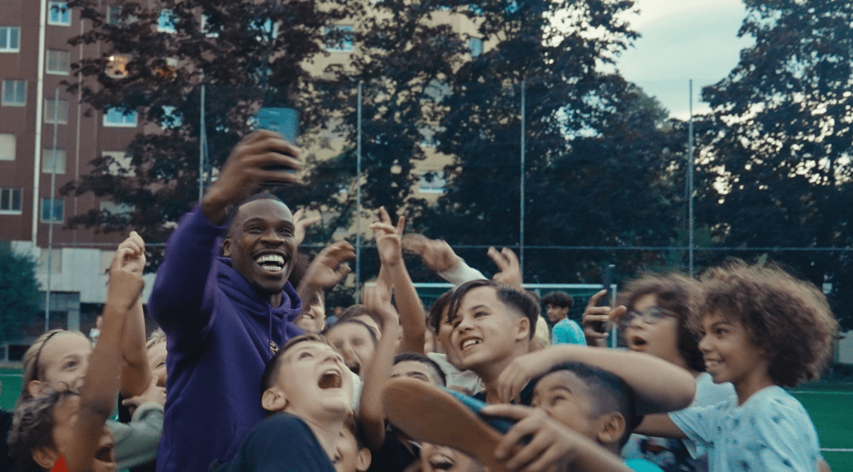 Un groupe d’enfants souriants prend un selfie avec un adulte sur un terrain de sport, entourés d’arbres et d’immeubles.