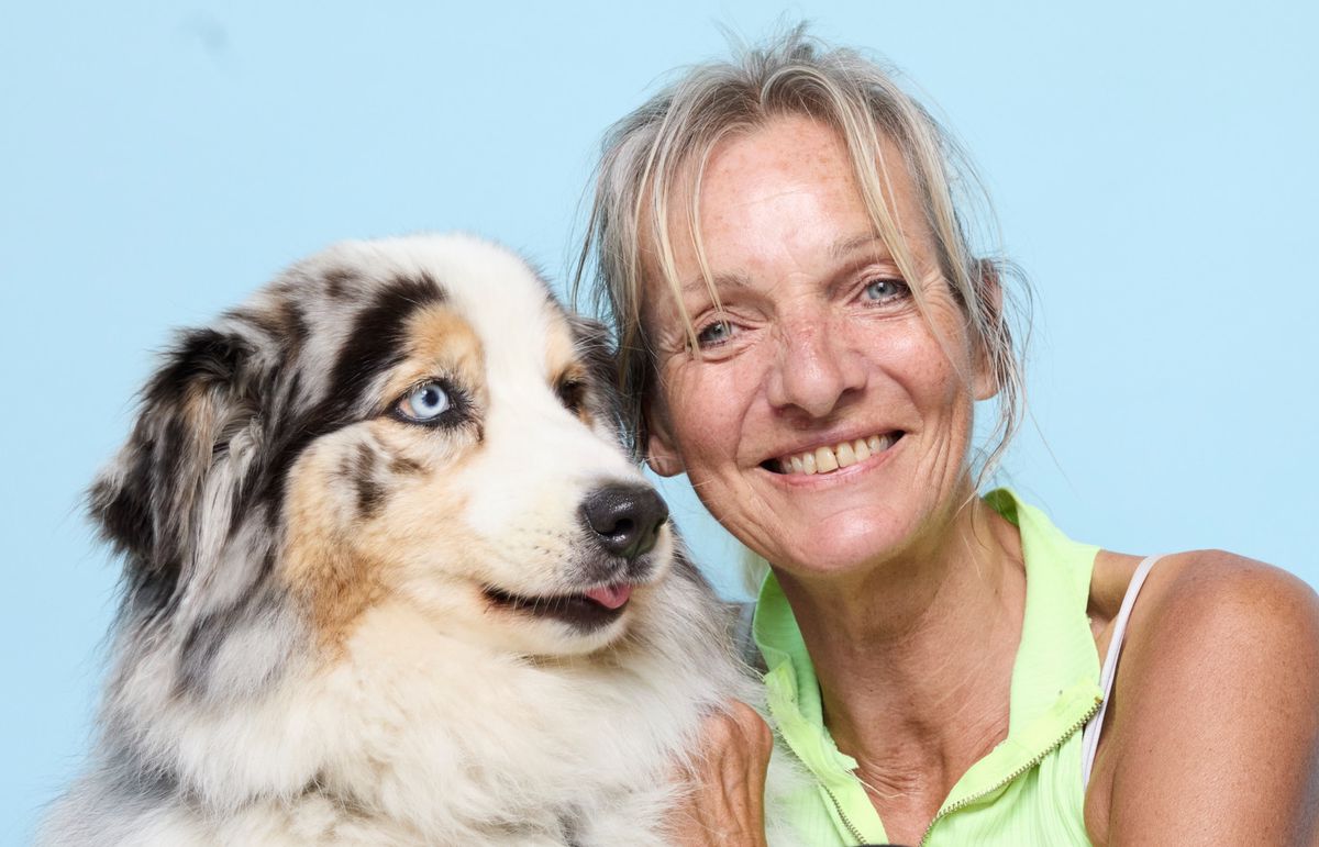Une femme souriante dans un débardeur vert clair étreint un grand chien à fourrure blanche et noire avec des yeux bleus, contre un fond bleu.