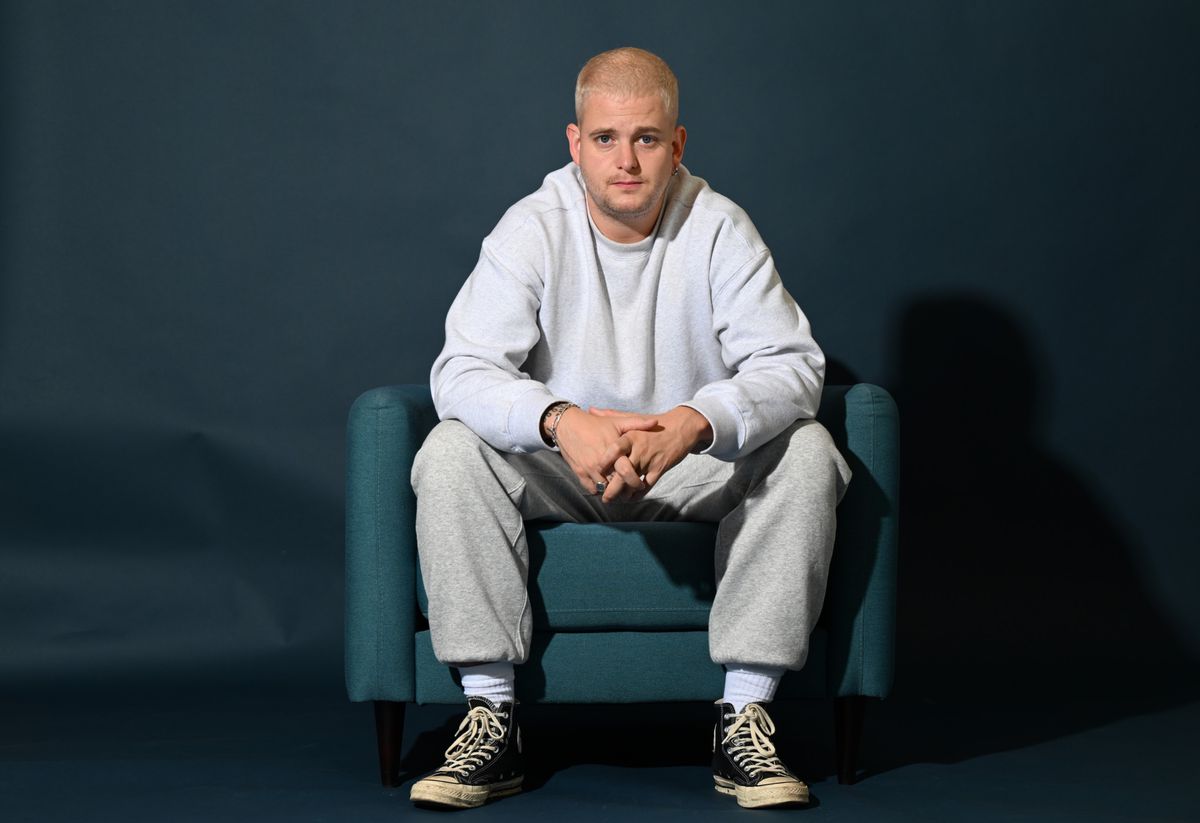 Ein Mann in grauem Freizeitanzug und schwarzen Turnschuhen sitzt auf einem blauen Sessel vor einem dunkelblauen Hintergrund.