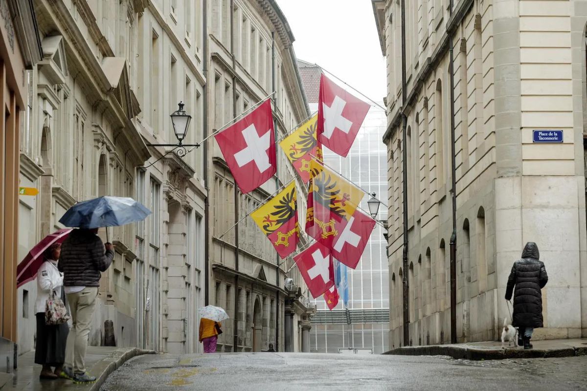 Rue de Genève sous la pluie avec des drapeaux suisses et genevois suspendus, des passants avec des parapluies.