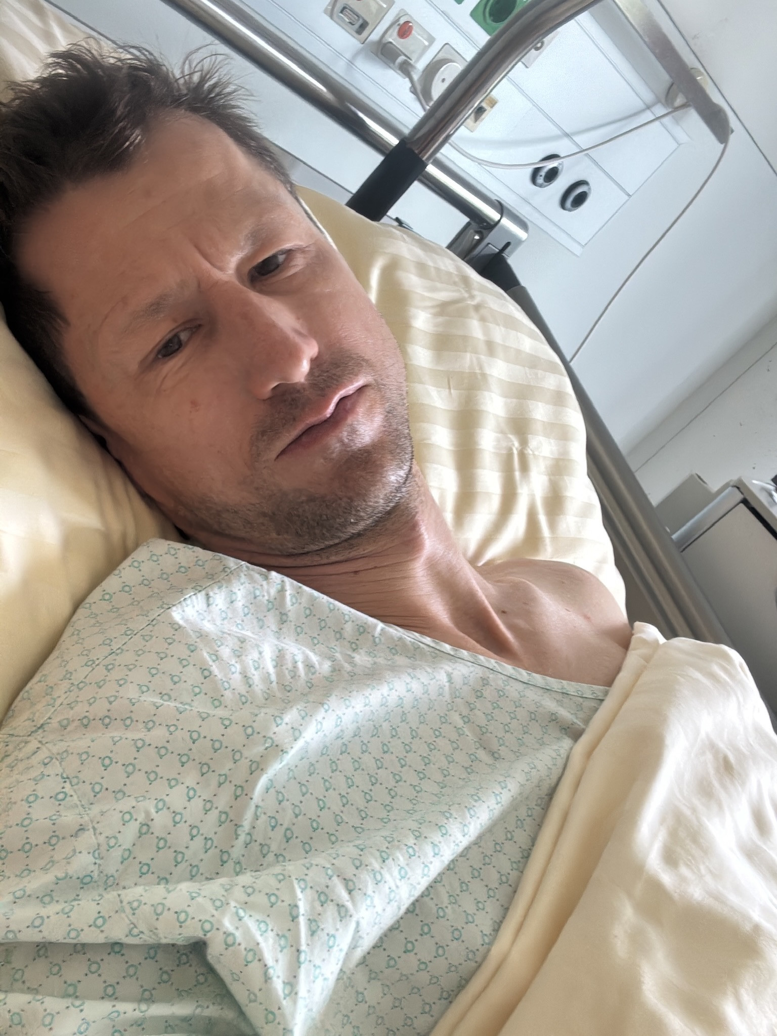 Mann liegt in einem Krankenhausbett mit Spitalkittel und blickt in die Kamera.