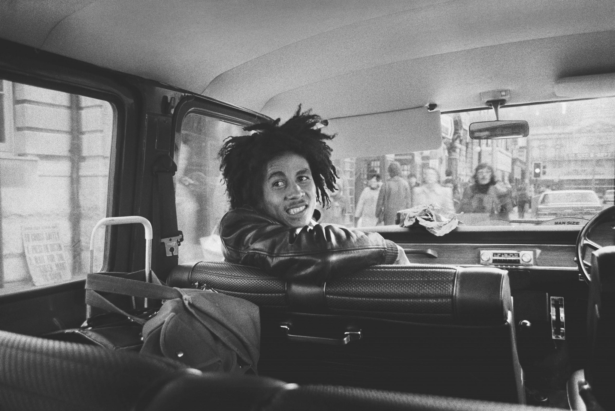 Une personne avec des cheveux coiffés en dreadlocks est assise à l’arrière d’une voiture, regardant en arrière avec un sourire, dans une scène urbaine animée.