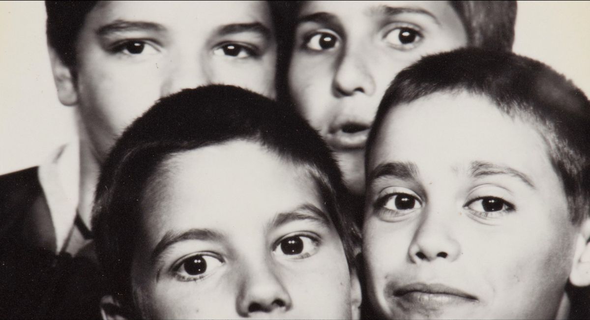 Quatre jeunes garçons posent pour une photo en noir et blanc, se serrant étroitement les uns contre les autres et regardant la caméra avec diverses expressions.