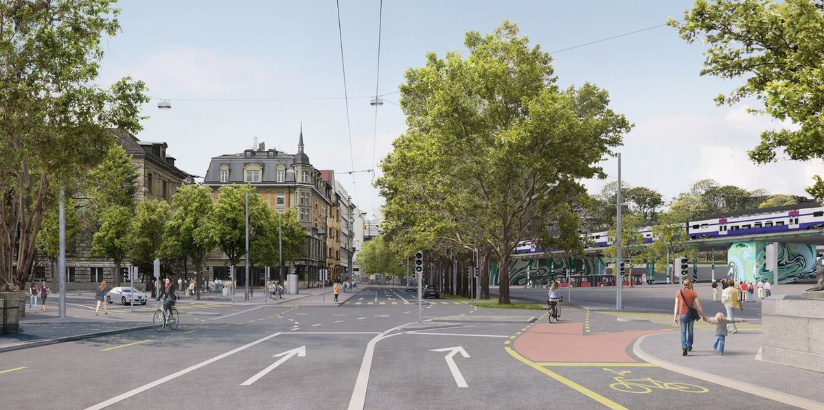 Kreuzung mit Rad- und Fusswegen, umgeben von Bäumen und Gebäuden mit einer Strassenbahnstation auf der rechten Seite, bei klarem Himmel.