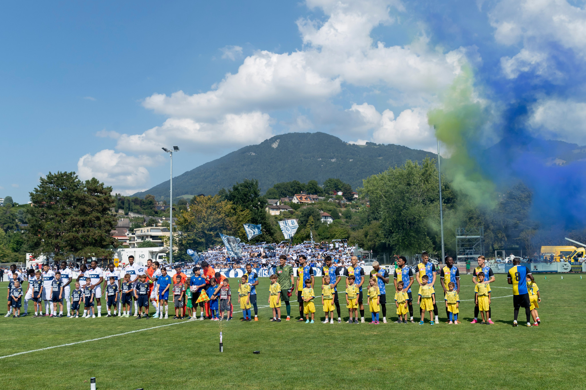 Vue des équipes de Vevey Sports et FC Lausanne-Sport alignées avant le match de la Coupe Suisse à La Tour-de-Peilz, avec des supporters et des fumigènes colorés en arrière-plan.