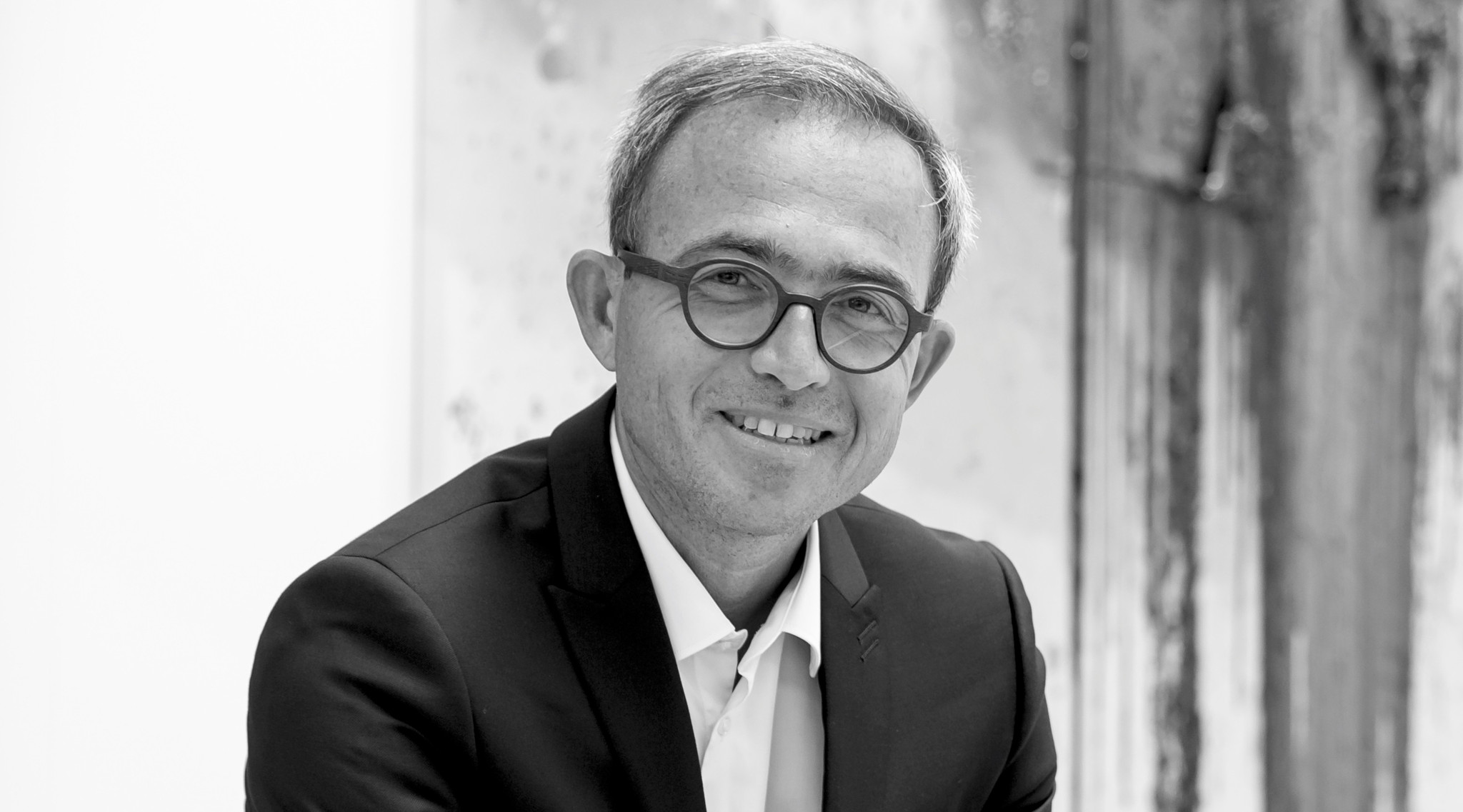 Black and white portrait of a smiling man wearing round glasses and a dark suit, in front of a light background.