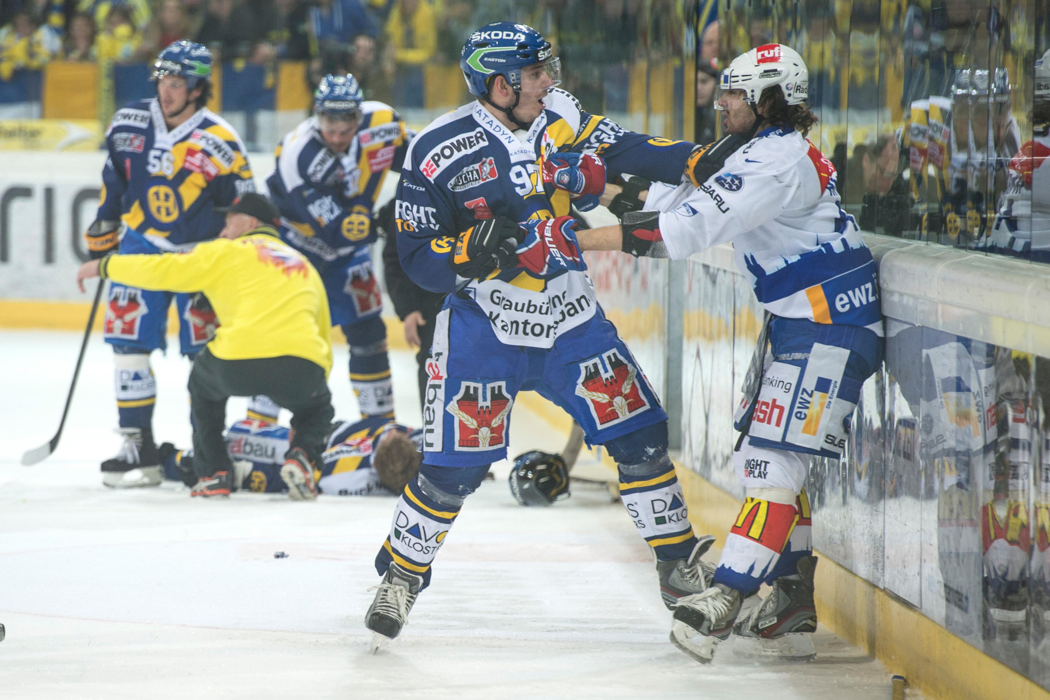 Eishockeyspieler in blauen Trikots kämpfen während eines intensiven Spiels an der Bande, im Hintergrund weitere Spieler und ein Schiedsrichter.