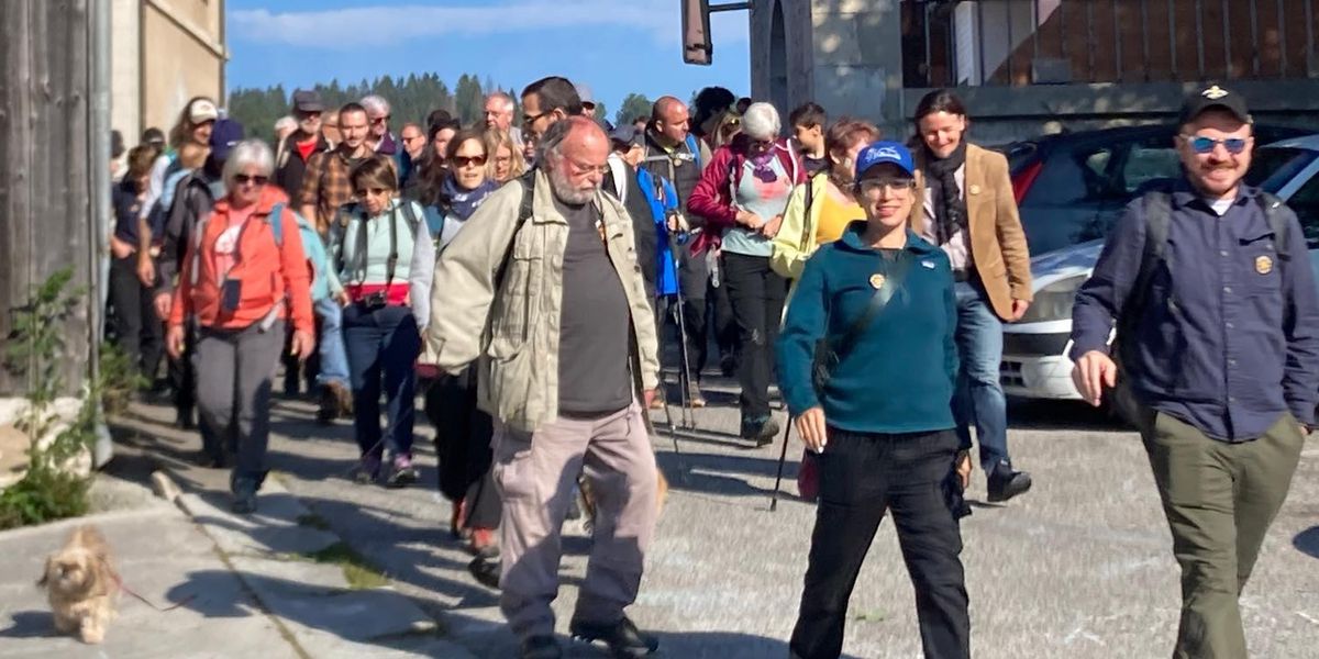 Un groupe de personnes marchant ensemble dans une rue, dans un village ensoleillé. Certains portent des sacs à dos, et il y a un petit chien sur le trottoir.