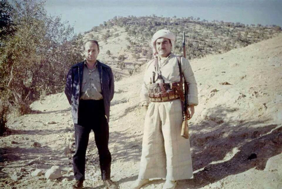 Deux hommes debout dans un paysage aride et rocheux, l&rsquo;un portant une tenue traditionnelle avec une ceinture de munitions et un fusil.
