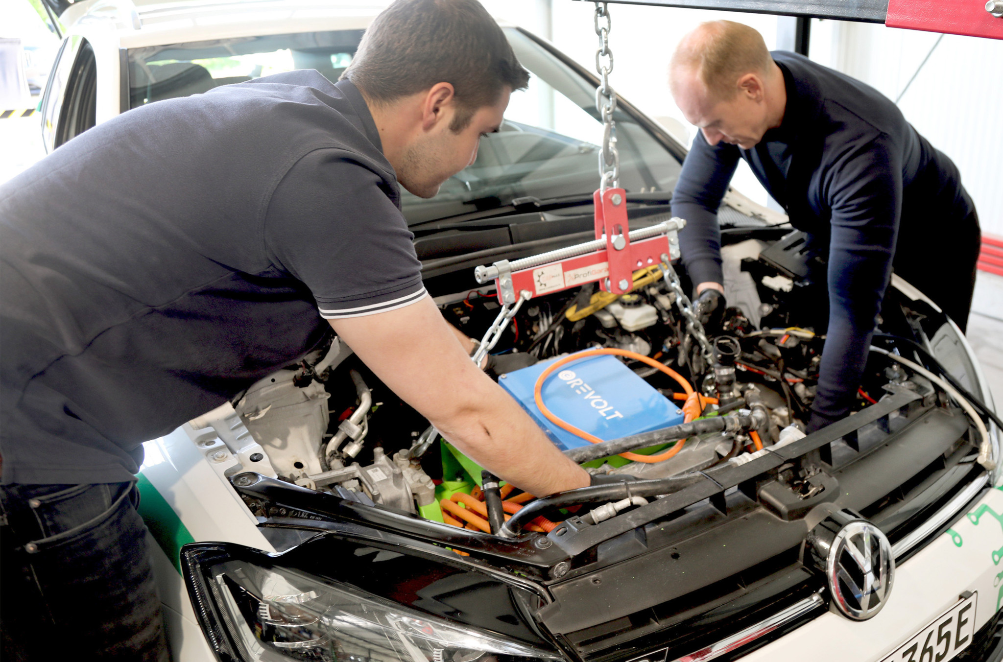 Zwei Techniker installieren einen Elektroantrieb mit blauer Batterie in einem VW-Fahrzeug in einer Werkstatt.