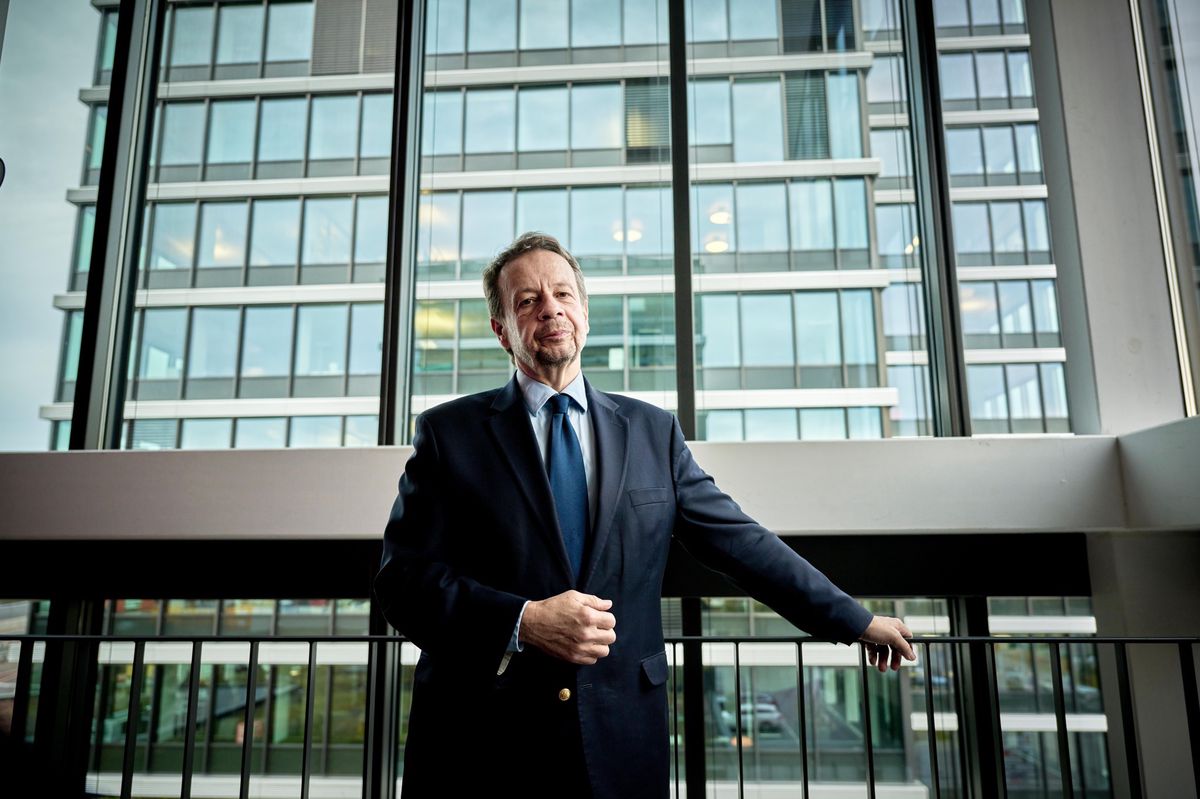 Un homme en costume se tient devant une fenêtre avec une vue sur un immeuble moderne, illustrant une interview avec le directeur général de la SRG.
