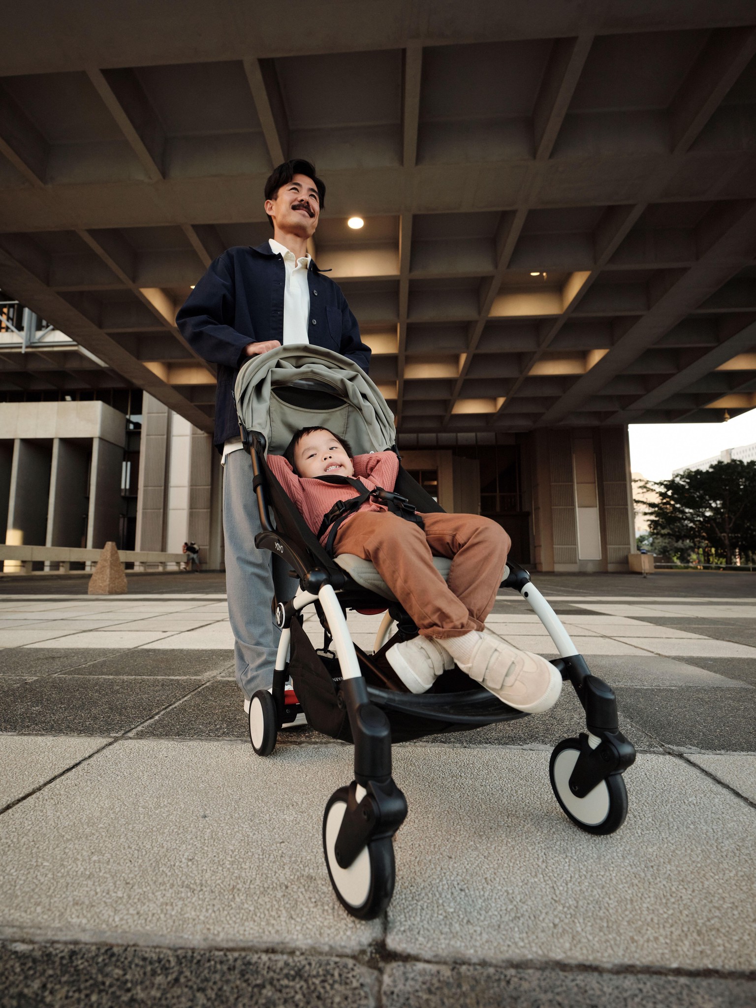 Un homme marche en ville avec un enfant dans le nouveau poussette Stokke YOYO³, illustrant le design urbain innovant du produit.