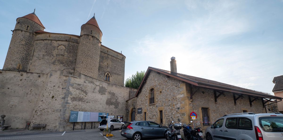 Bâtiment historique avec des tours en pierre et des toits en cône, à côté d'un bâtiment en pierre plus petit. Plusieurs véhicules, dont des voitures et des motos, garés devant.