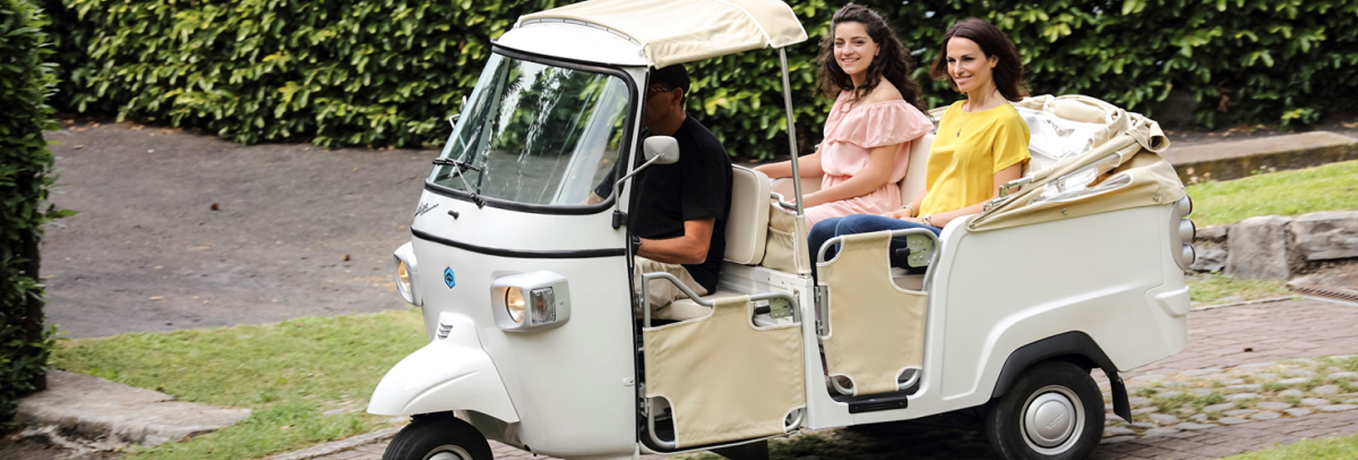 Ein weisses Tuk-Tuk fährt mit einem Fahrer und zwei Frauen als Passagiere auf einem gepflasterten Weg neben einer Hecke.