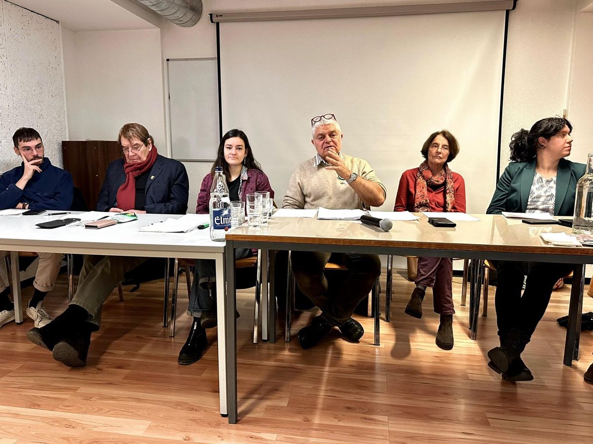 Sept personnes assises à une table lors d’une réunion, avec un micro devant elles, dans une salle avec un écran de projection.