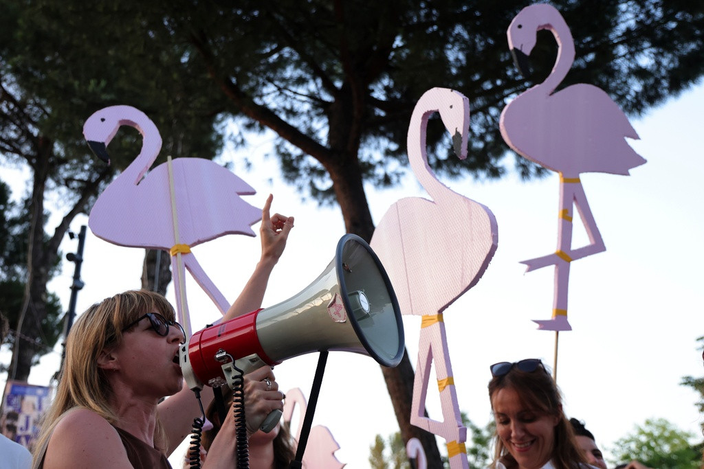 Demonstrantin mit Megafon an Protest in Tirana gegen Kushner-Küstenprojekt am Narta-Lagon, umgeben von Flamingo-Schildern.