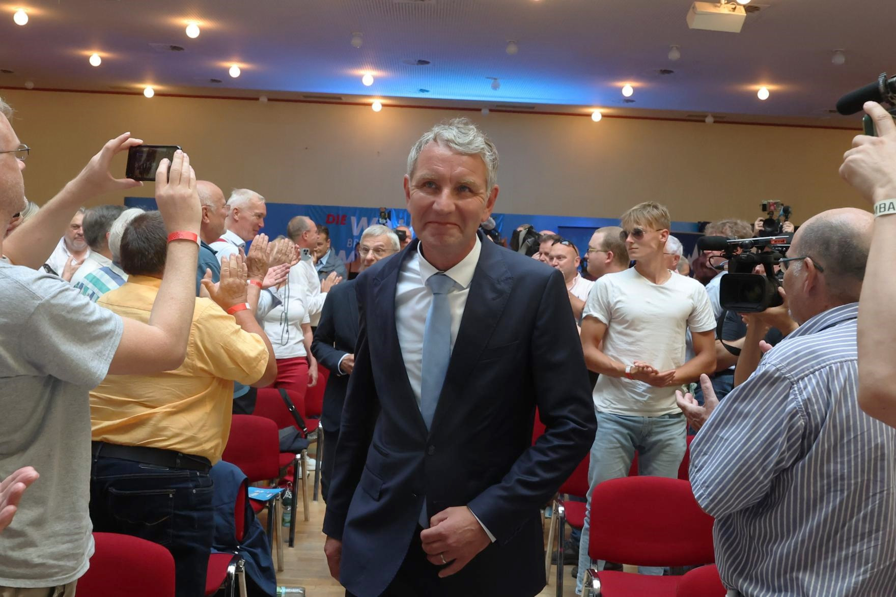 Ein Mann in einem Anzug steht in einem Raum voller Menschen, von denen einige Fotos machen. Es gibt rote Stühle im Hintergrund.