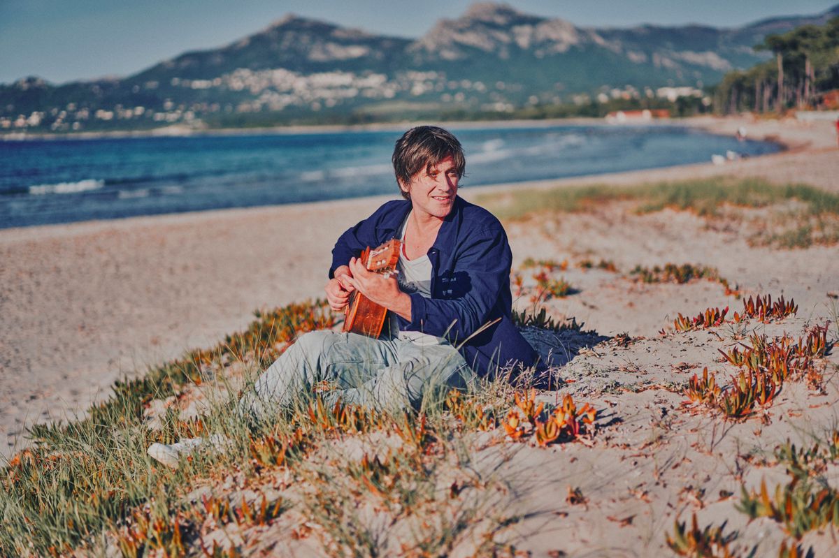 Thomas Dutronc jouant de la guitare assis sur une plage avec des montagnes en arrière-plan et la mer à gauche.