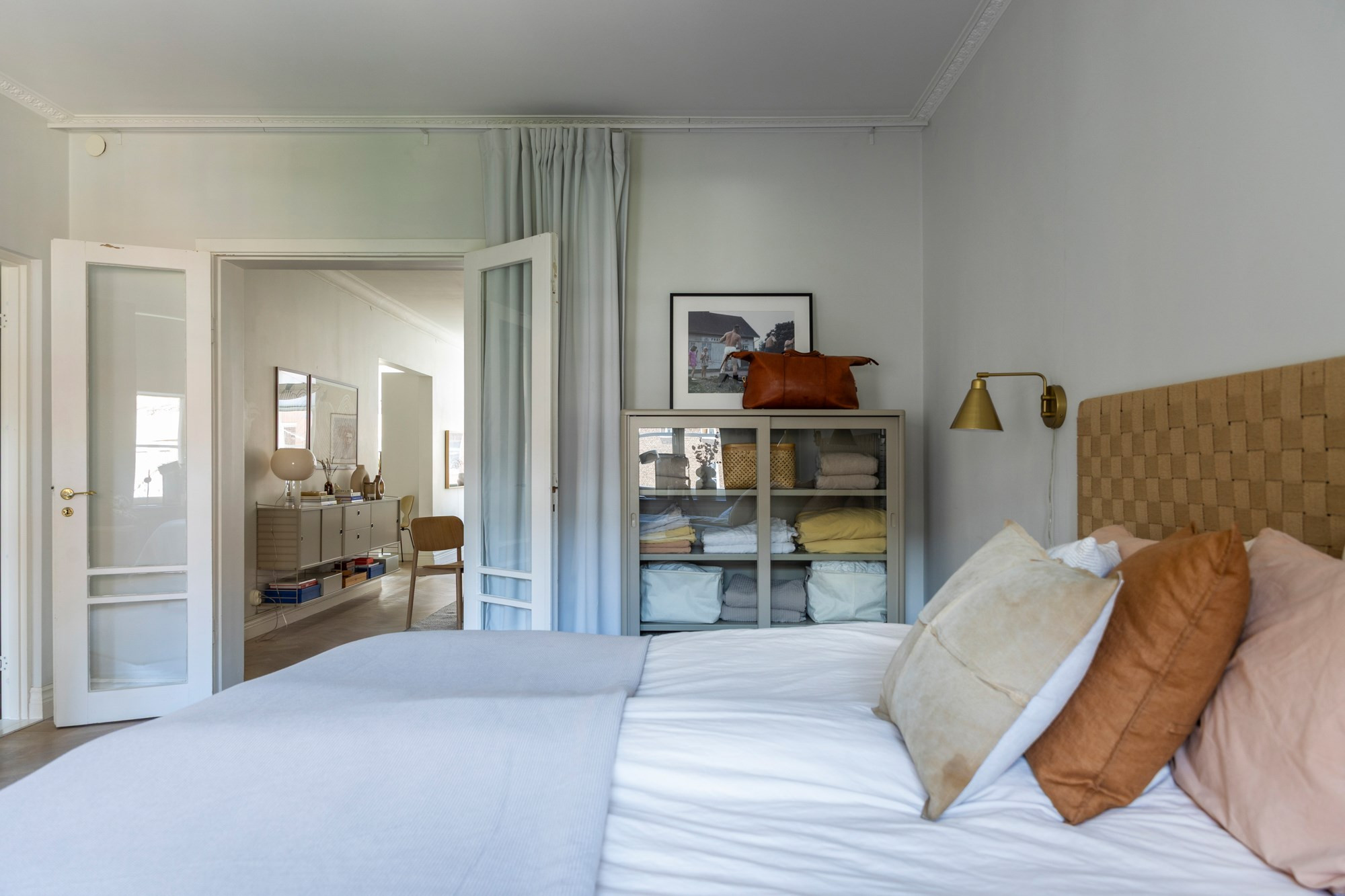 Helles Schlafzimmer mit Doppelbett, geflochtenem Kopfteil, bunten Kissen, Regalschrank und offener Tür zum Nebenraum.