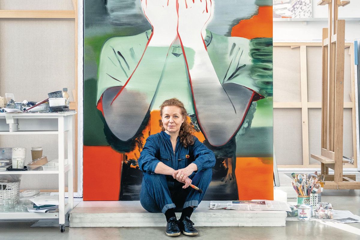 Une femme assise en combinaison bleue devant une grande peinture représentant une personne se couvrant le visage avec ses mains dans un atelier d’art.