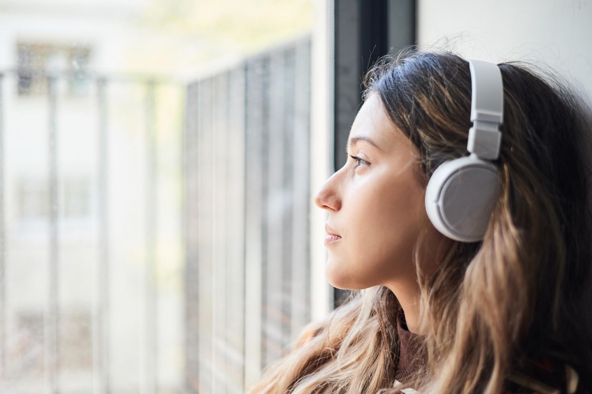 Jeune femme triste et stressée écoutant de la musique en regardant par la fenêtre, illustrant le concept de santé mentale.