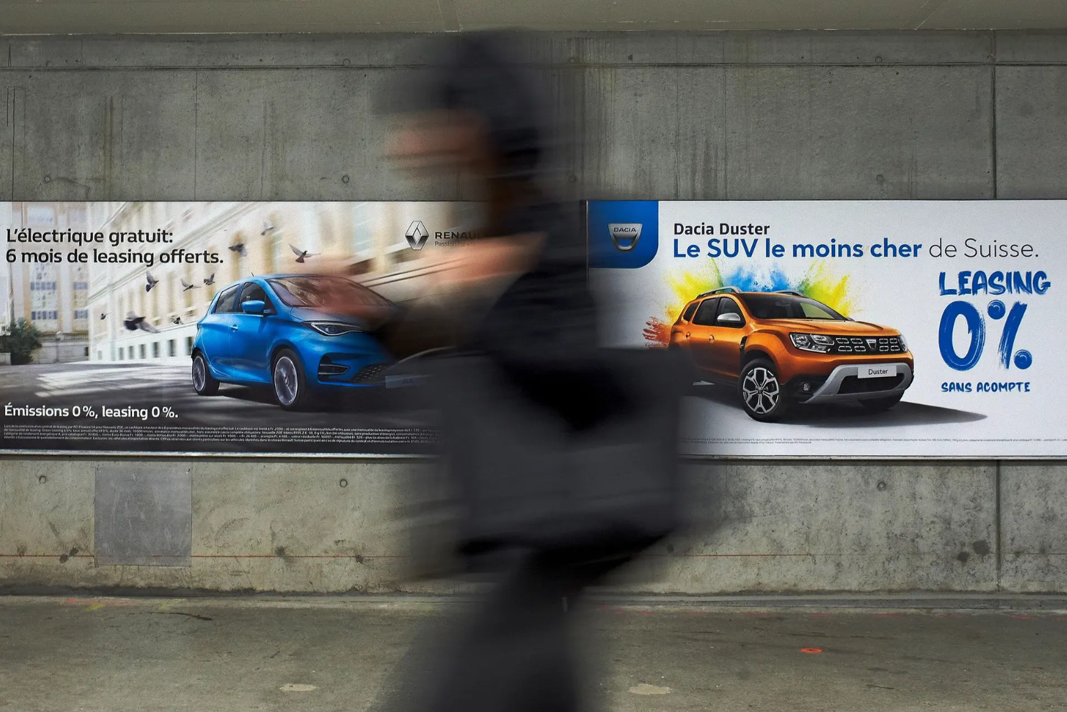 Personne floue passant devant des affiches publicitaires pour des voitures Renault et Dacia, mettant en avant des offres de leasing.