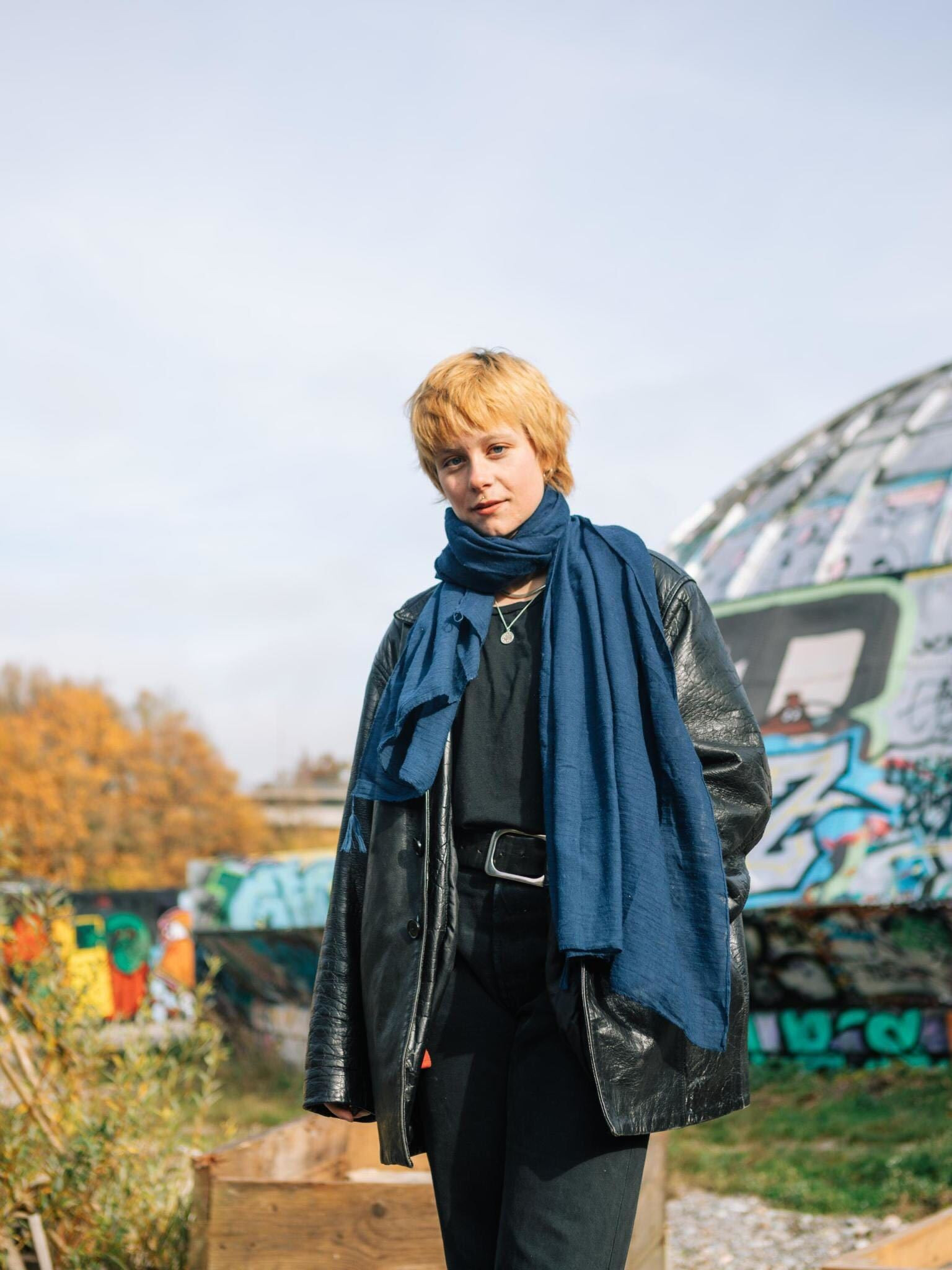 Eine Frau mit blonden Haaren steht vor graffiti-bedeckten Strukturen, trägt eine schwarze Lederjacke und einen blauen Schal. Im Hintergrund sind bunte Herbstbäume zu sehen.