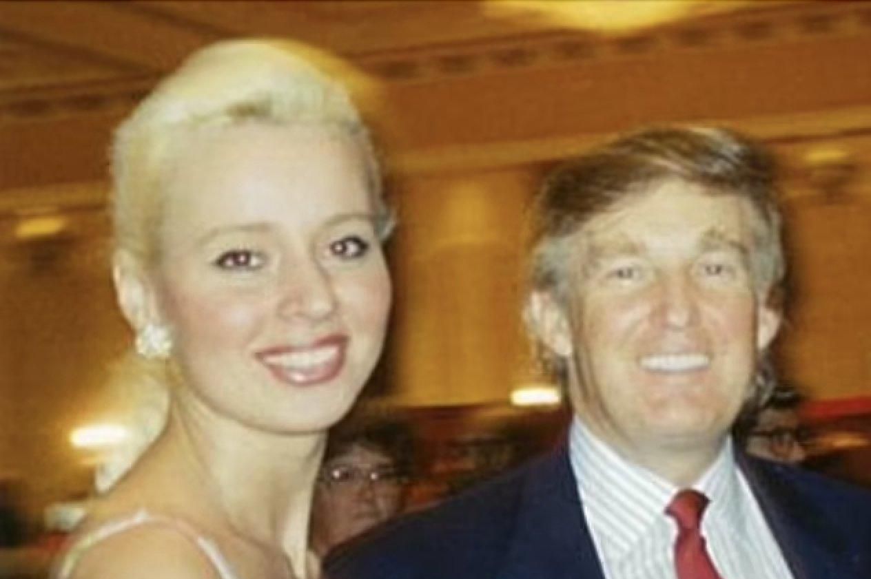 Un homme en costume et une femme en robe florale, dans un environnement intérieur éclairé.