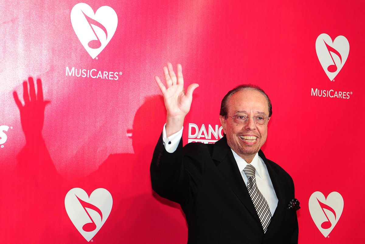 Le musicien brésilien Sergio Mendes salue à son arrivée au Tribute MusiCares Person of the Year 2012 en l'honneur de Paul McCartney à Los Angeles. Mendes est vêtu d'un costume noir sur un fond rouge avec des logos MusiCares.