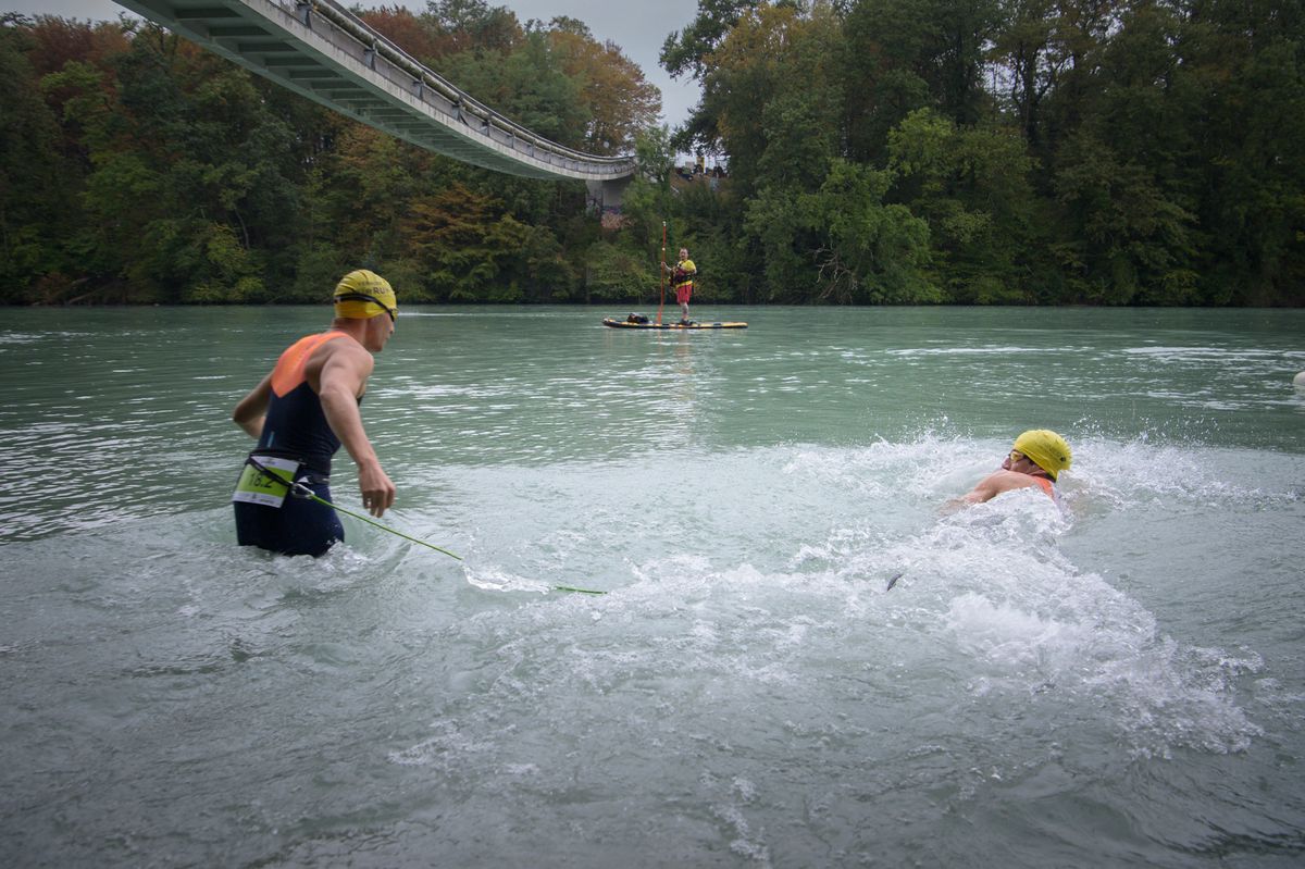 Deux nageurs portant des bonnets de bain jaunes traversent une rivière sous une passerelle, avec un bateau de sauvetage à l'horizon.