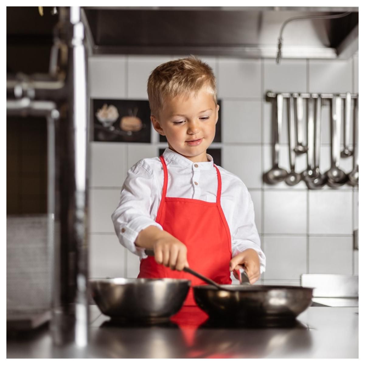 Un jeune garçon en tablier rouge cuisine dans une cuisine professionnelle, remuant une poêle tout en tenant un bol.
