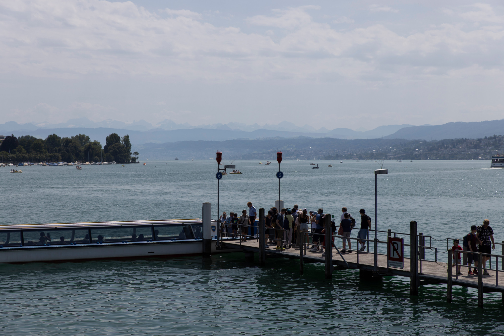 Menschen warten am Schiffssteg beim Bürkliplatz auf ein Boot, im Hintergrund der Zürichsee mit Alpenkulisse.