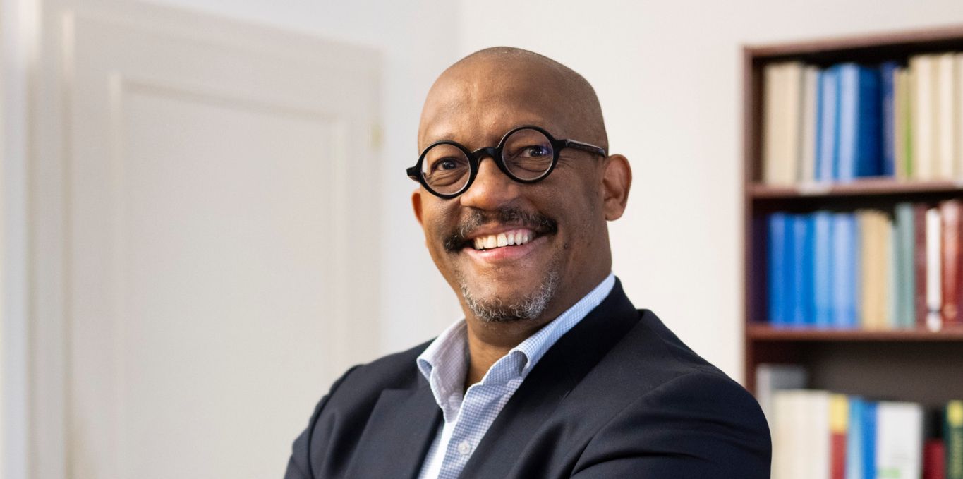Homme souriant avec des lunettes rondes et un costume, les bras croisés, appuyé contre une table en bois. Bibliothèque avec des livres en arrière-plan.