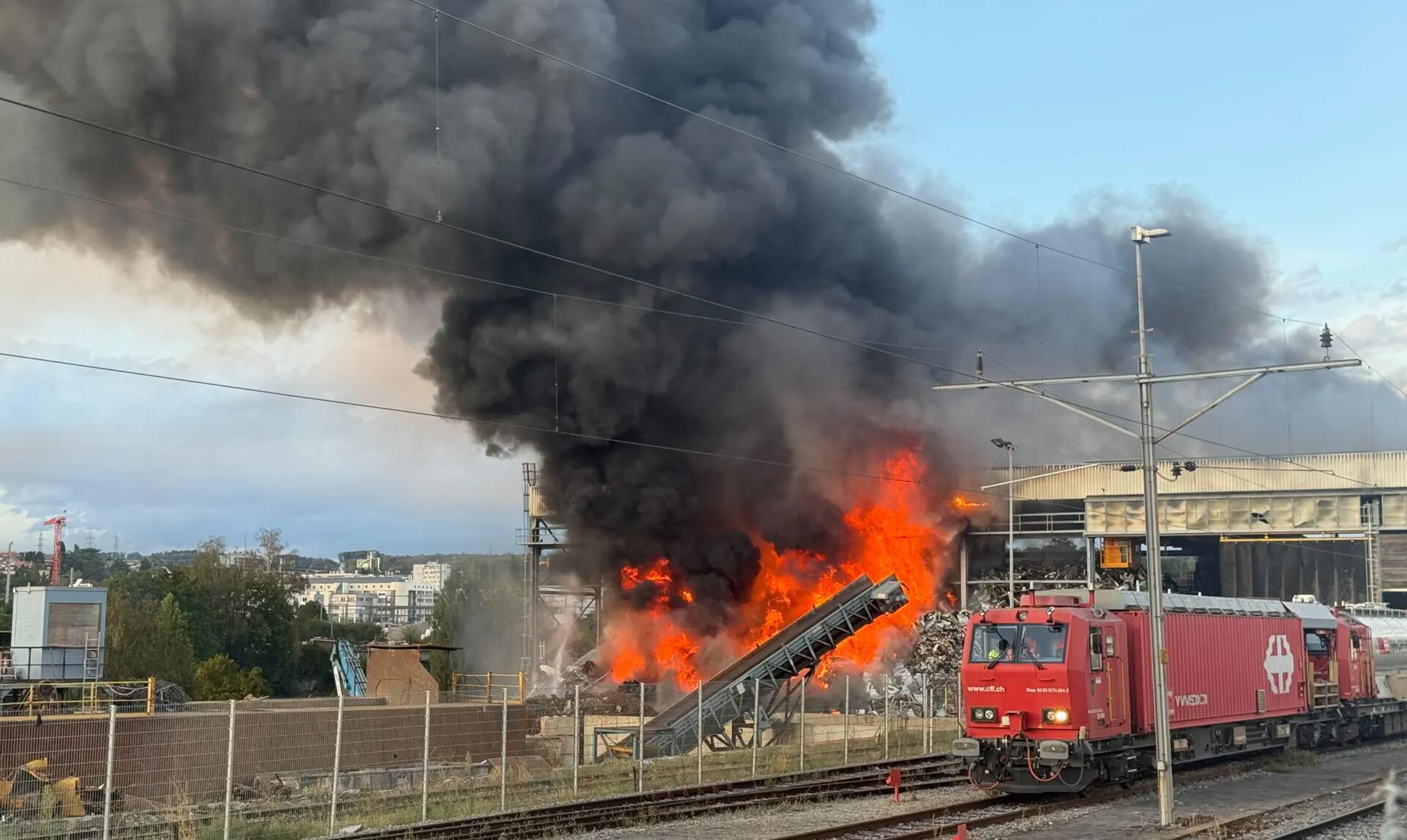 Grossbrand auf einem Bahngelände mit meterhohen Flammen und dichtem schwarzem Rauch, daneben eine rote SBB-Lokomotive.