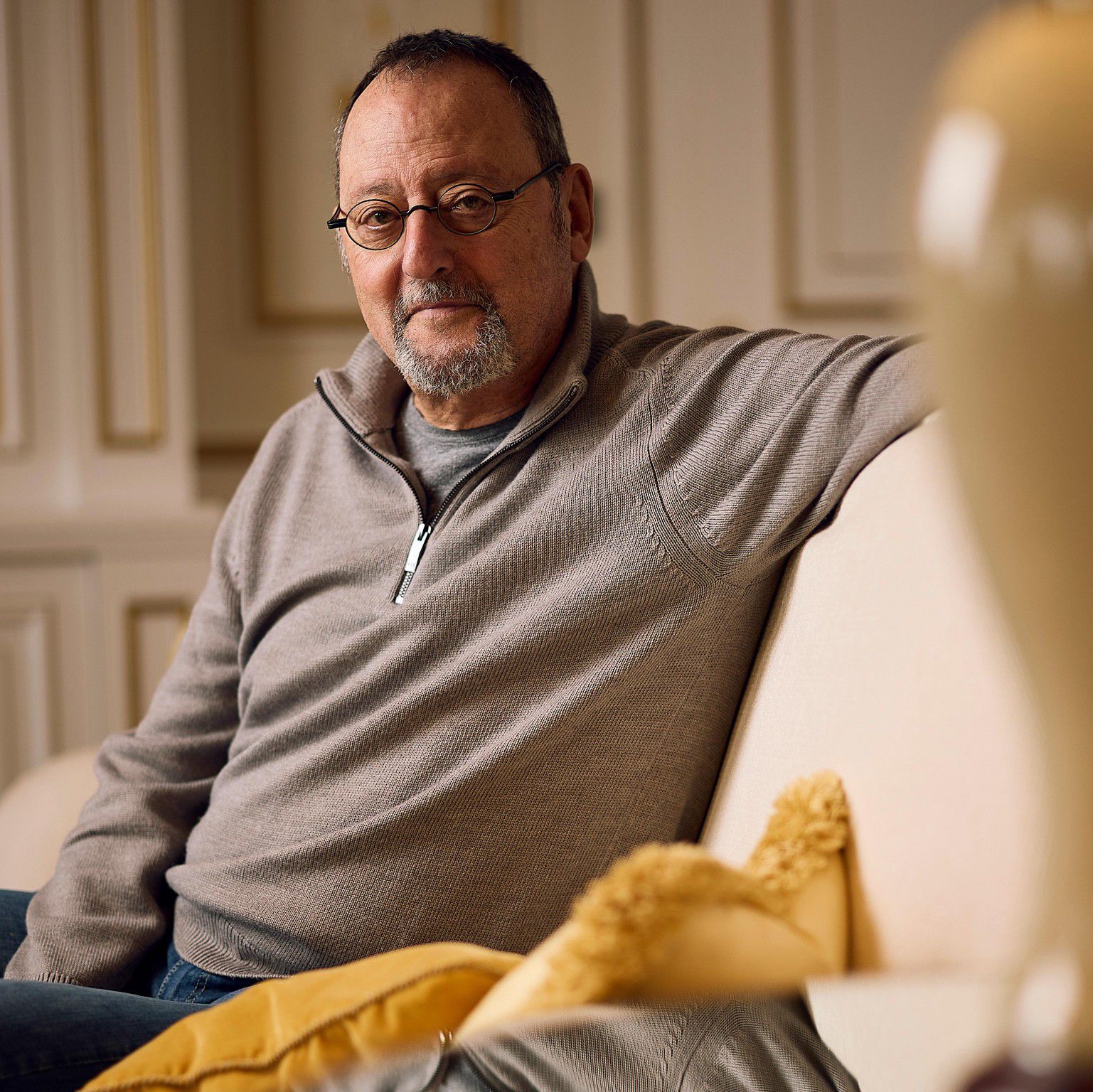 Jean Reno assis sur un canapé beige avec des coussins jaunes, reflété sur une table en verre.