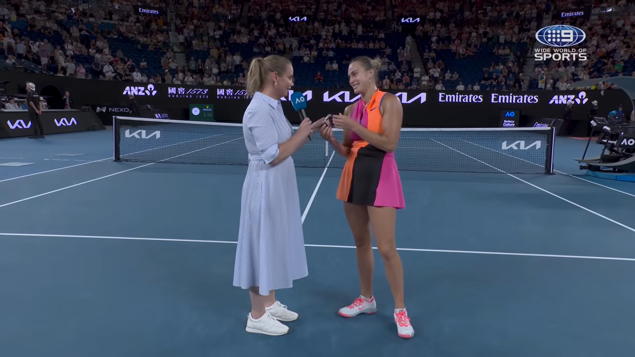 Two women stand on a tennis court while talking. One is wearing a blue dress and sneakers, the other is wearing a sporty outfit in orange and black. Spectators can be seen in the background.