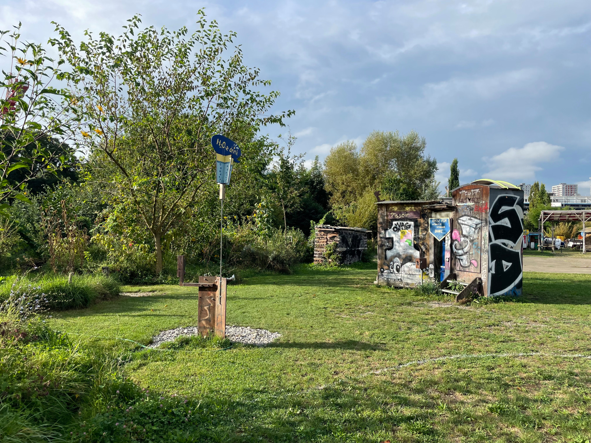 Ein gepflegter Garten mit Hochbeeten und Sonnenblumen im Vordergrund. Im hinteren Bereich des Gartens steht ein Tisch mit Stühlen unter einem kleinen Pavillon. Die Umgebung ist grün und von Bäumen umgeben.