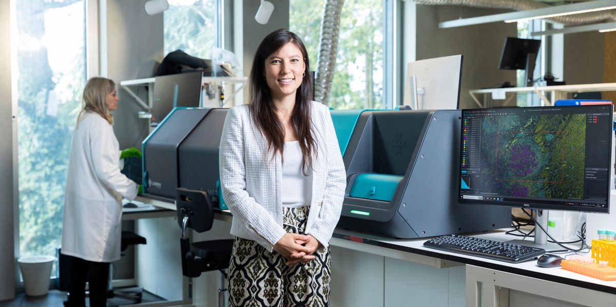 Scientifique souriante posant dans un laboratoire avec un collègue en blouse blanche travaillant à l'arrière-plan.