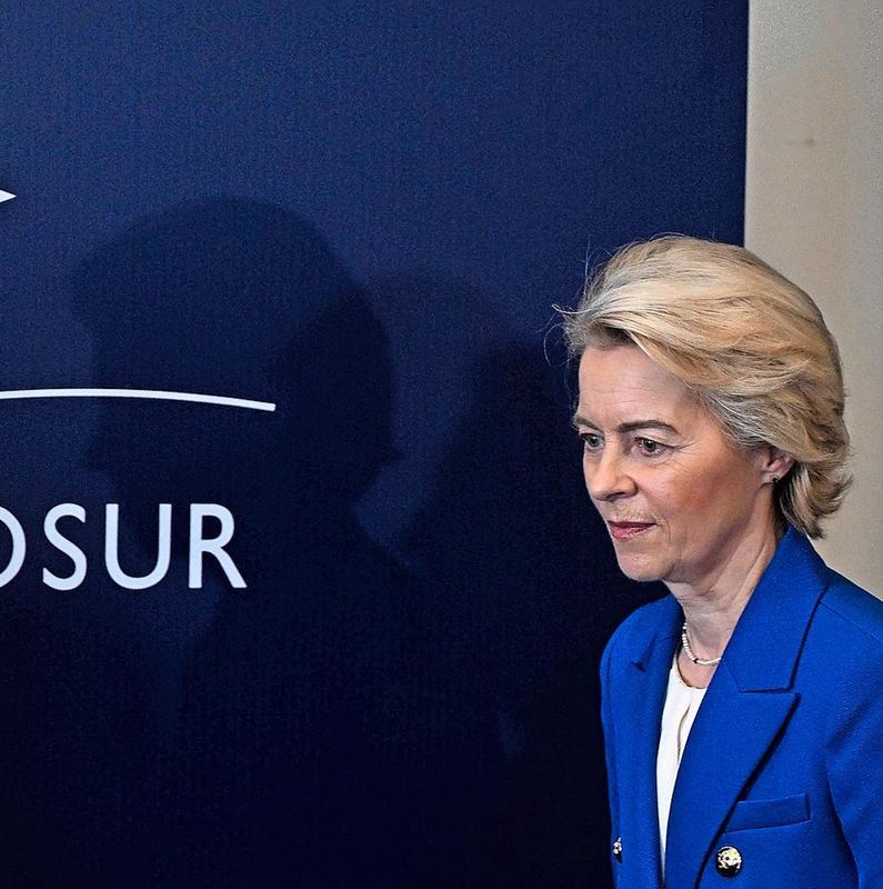 Femme en costume bleu devant un panneau bleu avec le texte ’MERCOSUR’ et étoiles blanches.