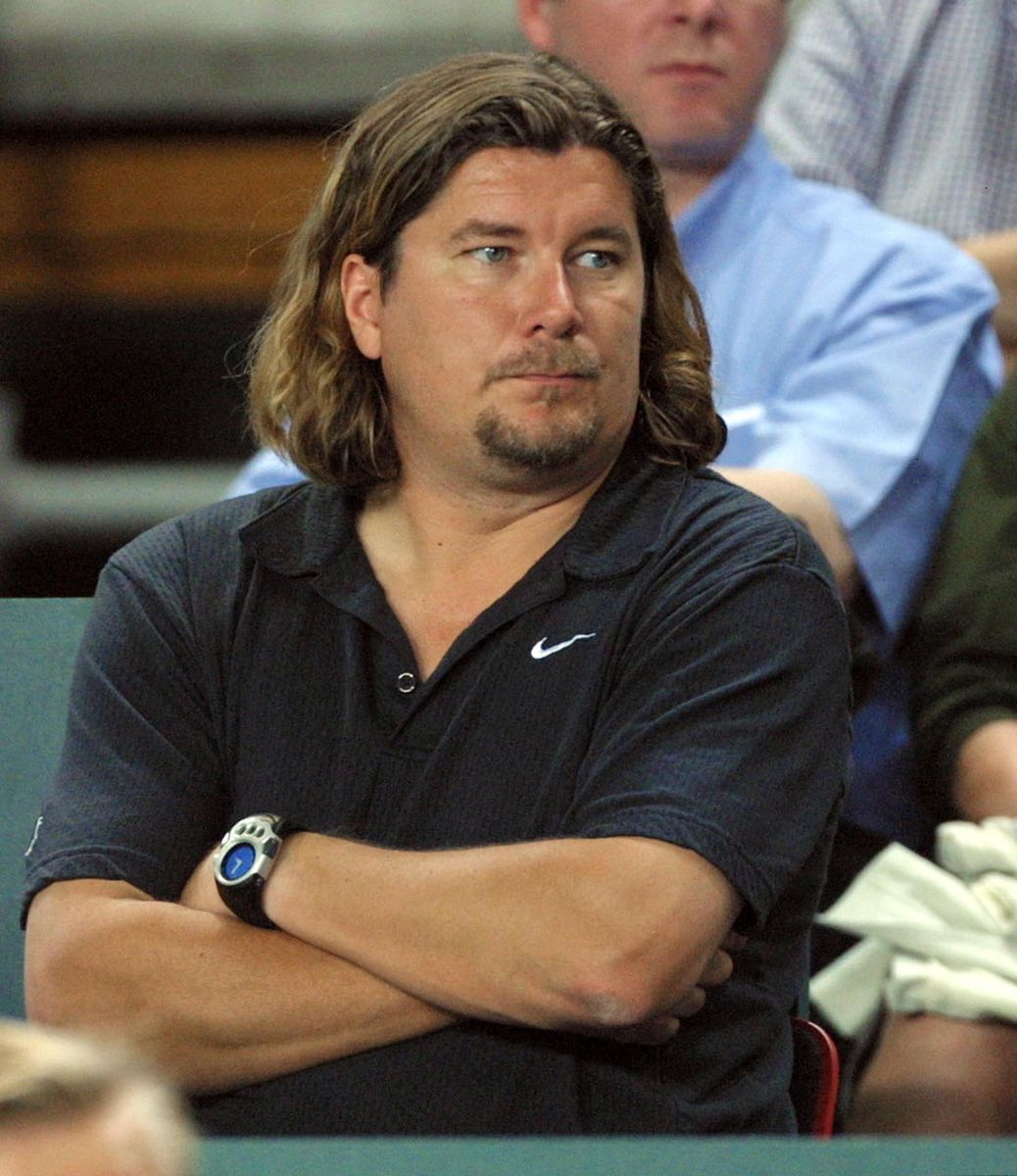 Un homme avec les cheveux longs et une barbe, vêtu d'un polo Nike, regarde quelque chose hors du cadre avec les bras croisés.